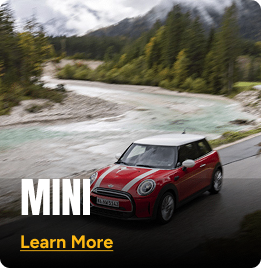 Red Mini Cooper driving alongside a river road with mountains and trees in the background. Click to learn more